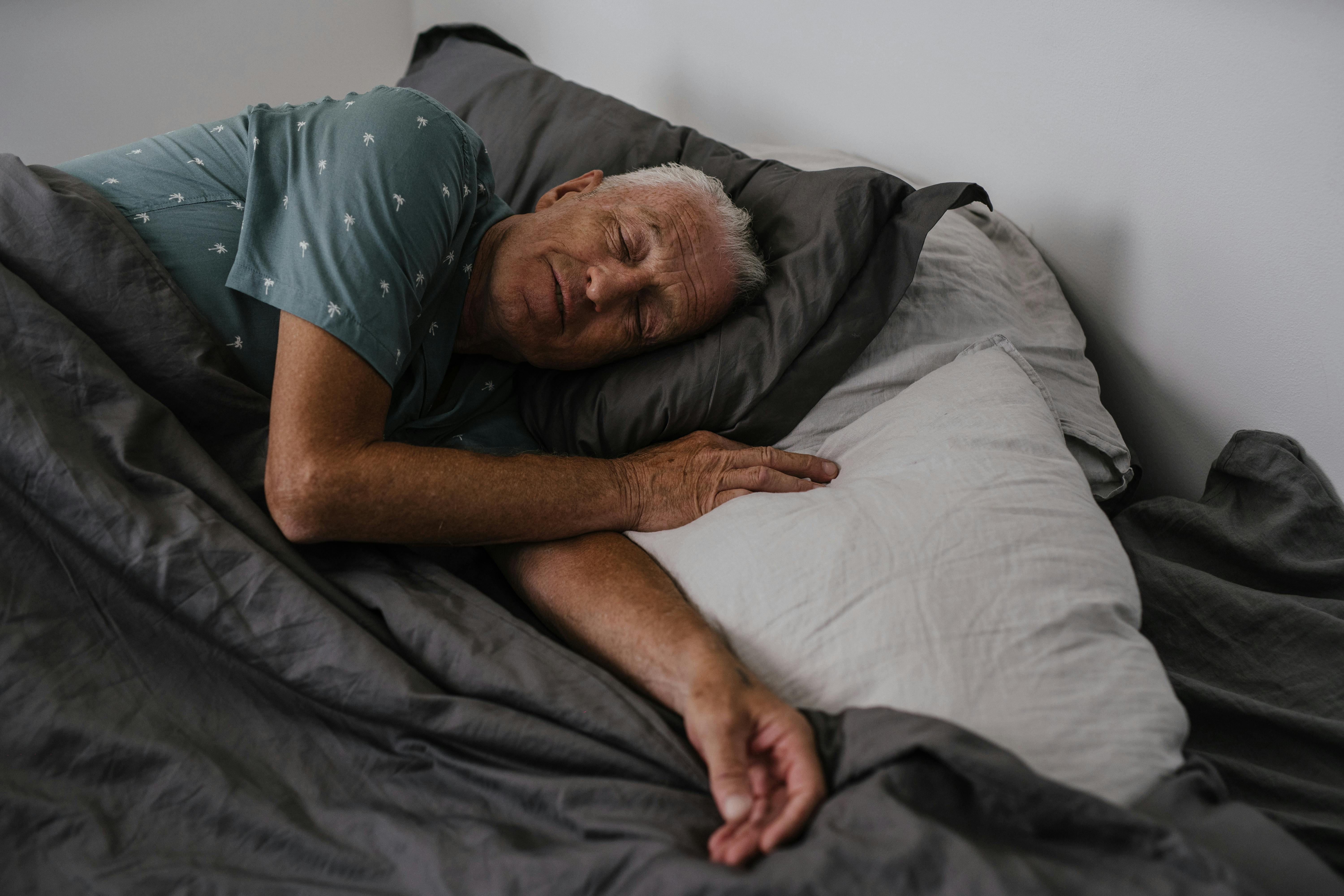 Old man sleeping peacefully in bed with gray bedding and a white pillow.
